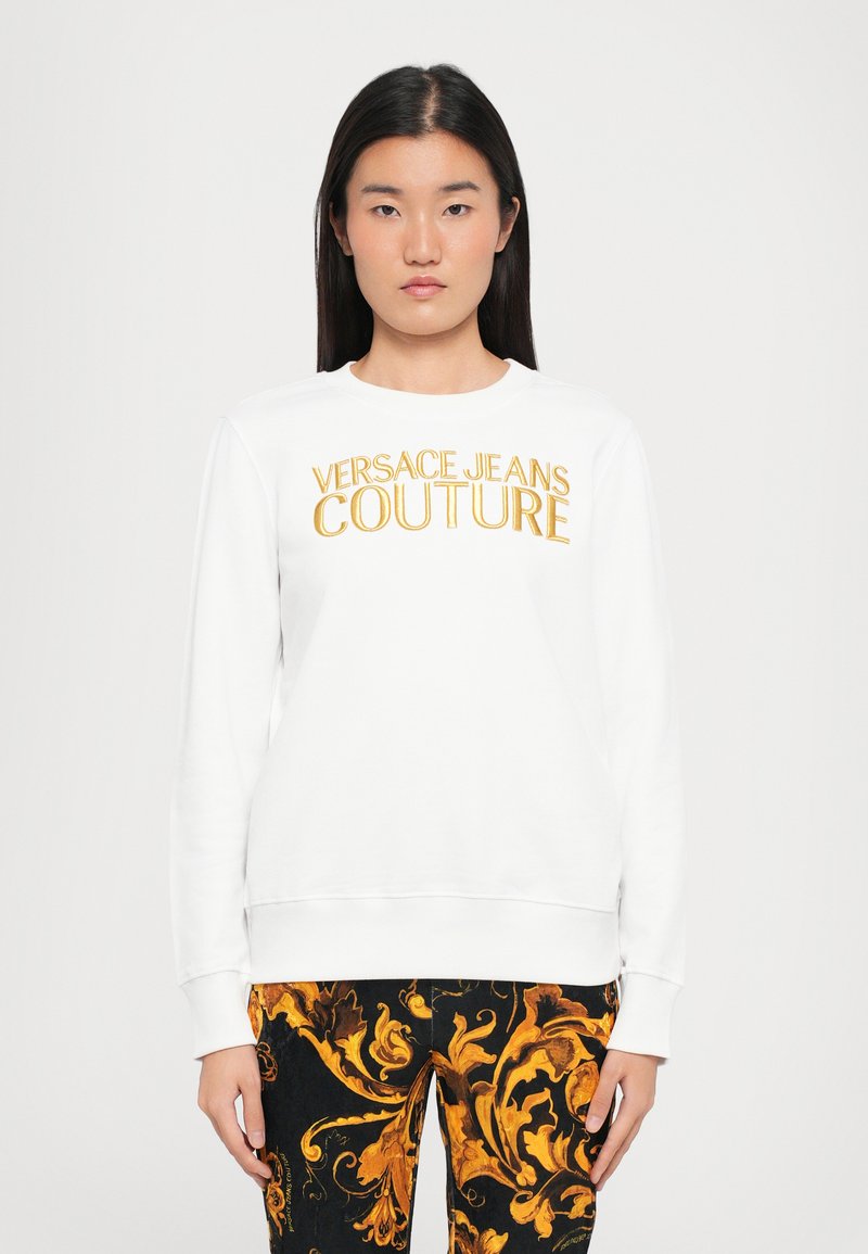 Felpa bianca con "VERSACE JEANS COUTURE" in lettering dorato. Polsini e orlo a coste, abbinata a pantaloni neri e dorati a fantasia.