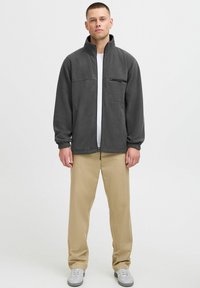 Veste en polaire grise avec col haut, fermeture éclair à l'avant et deux poches latérales. Comprend une poche poitrine zippée et des poignets élastiques.