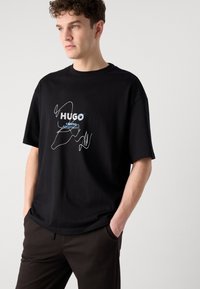 Black cotton t-shirt with short sleeves, featuring white and blue graphic text “HUGO” and abstract line art on the front.