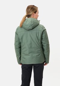 Giacca isolante verde con cappuccio, caratterizzata da una superficie liscia, cuciture sul retro e orlo corto. Indossata con pantaloni neri.