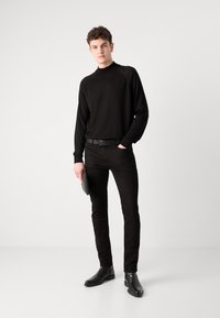 Pull à col roulé noir à manches longues, pantalon noir slim, ceinture noire et bottines noires. Sujet tenant une pochette noire dans la main gauche.