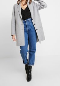 Cappotto di lana grigio con rever a punta, bottoni neri e vestibilità rilassata, abbinato a jeans blu a vita alta e stivaletti neri.
