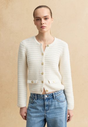 Donna con capelli biondi legati indossa un cardigan bianco testurizzato con bottoni dorati e jeans blu a vita alta su sfondo beige.