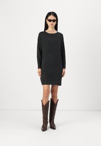 Robe pull en maille gris charbon avec des manches longues, un col rond et un ourlet inégal. Associée à des bottes de cowboy en cuir marron jusqu'aux genoux.