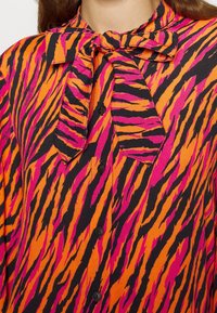 Langemouwen blouse in een levendige oranje, roze en zwarte tijgerstreep patroon. Bevat een strik-sluiting kraag en knopen aan de voorkant.
