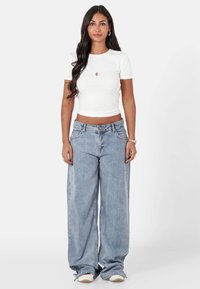 Camiseta blanca corta con un pequeño logo rojo, combinada con jeans de pierna ancha en azul claro con cintura alta y textura clásica de denim.