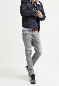 Man som bär en mörk denimjacka, lager med huvtröjor, ljusgrå jeans och svarta sneakers, stående med benen korsade mot en neutral bakgrund.