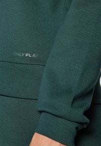 Tissu de sweat-shirt vert foncé avec une texture lisse ; présente une couture horizontale et le texte "ONLY PLAY" en argent au bas.
