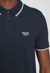 Polo bleu marine avec un col classique, trois boutons, et des accents à rayures blanches sur le col. Présente un logo blanc brodé bien visible.