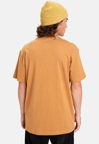 Bruine katoenen t-shirt met korte mouwen en een relaxte pasvorm, gedragen met een gele gebreide beanie. Zachte textuur, eenvoudig ontwerp, geen zichtbare logo's.