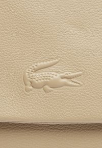 Texture en cuir beige avec un logo de crocodile en relief bien en vue. La surface est légèrement texturée, offrant une apparence douce et souple.
