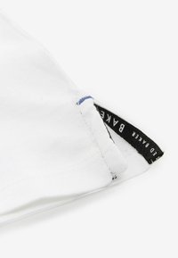 Camiseta blanca de algodón con cuello redondo, que presenta acentos de costura azul y una etiqueta negra con texto blanco en el dobladillo.