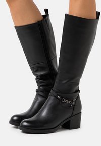Botas de cuero negro hasta la rodilla con tacón cuadrado, que cuentan con un cierre lateral de cremallera y una correa con un detalle de cadena metálica. Textura suave y diseño estructurado.