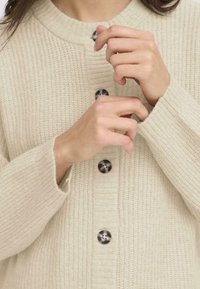 Šviesiai smėlinis nertinis kardiganas su tekstūruota paviršiumi, su dideliais tamsiais mygtukais ir apykakle. Rankos taiso apykaklę.