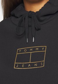 Sudadera negra con el logotipo dorado de Tommy Jeans en el pecho y cordones negros con el nombre de la marca en los terminales de metal.