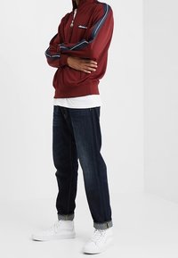 Persoon in een bordeauxrode ritsjack met marineblauwe en witte strepen, donkere opgerolde spijkerbroek en witte hoge sneakers, staand met de armen gekruist.