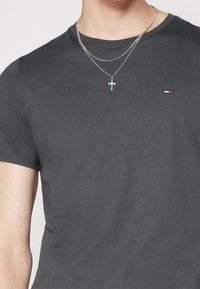Grau Baumwoll-T-Shirt mit kurzen Ärmeln, verfügt über ein kleines farblich abgesetztes Logo auf der Brust und geschichtete silberne Halsketten mit einem Kreuzanhänger.