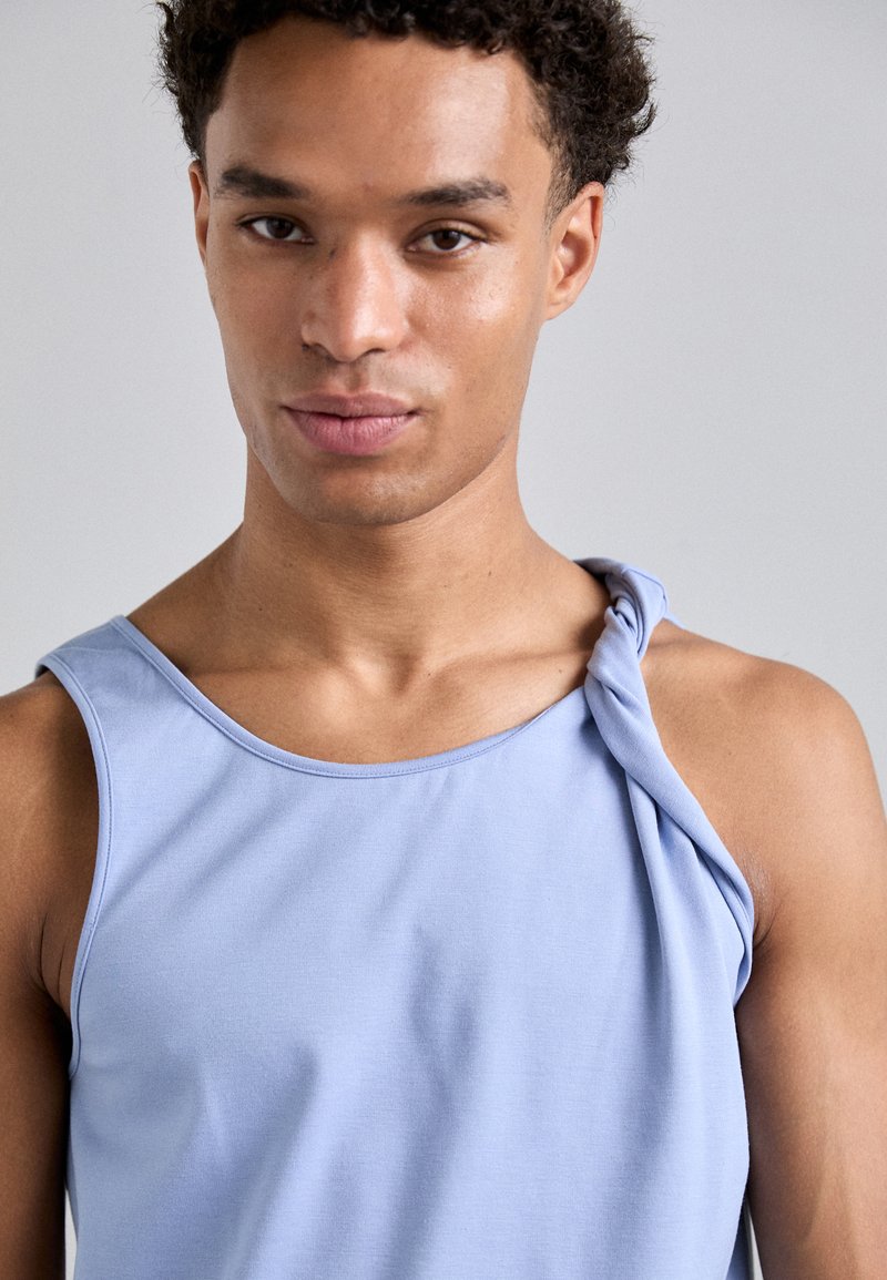 Lichtblauw mouwloos topje met een gedrapeerde knoopdetail op één schouder, gemaakt van soepel, rekbaar materiaal, met een ronde halslijn.