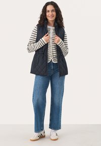 Marineblau gestepptes Weste mit hohem Kragen und Zugband in der Taille, getragen über einem gestreiften Langarmshirt. Kombiniert mit weit geschnittenen blauen Jeans und Sneakers.