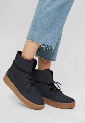 Baskets montantes en daim bleu marine avec des lacets noirs et une semelle en caoutchouc marron. Ourlet effiloché sur un jean bleu clair visible au-dessus des chaussures.