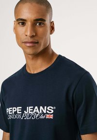 Navyblauwe katoenen t-shirt met witte tekst "PEPE JEANS LONDON PJL73Club" en een klein Union Jack-logo. Korte mouwen, ronde halslijn.