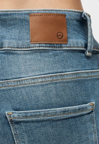 Κοντινή λήψη της πίσω τσέπης και της μέσης του μπλε υφασμάτινου τζιν, με ραμμένο καφέ δερμάτινο ταμπελάκι που φέρει ένα μικρό κυκλικό λογότυπο.