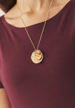 Közeli kép egy arany medál nyakláncról, amely három texturált kör alakú koronggal van díszítve, és lila felső fölött viselik.