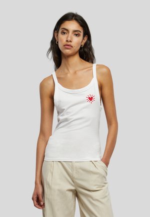 Femme aux longs cheveux foncés porte un débardeur blanc avec un motif cœur rouge sur la poitrine et un pantalon beige clair sur fond gris.