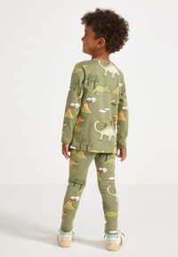 Ragazzo in piedi di spalle, che indossa un pigiama verde con stampe colorate di dinosauri e vulcani e sneakers bianche con calzini blu.