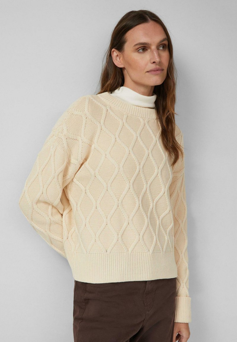 Cremfarbener Pullover mit einem Rautenmuster, gerippten Bündchen und Saum, im lässigen Schnitt über einem weißen Rollkragenpullover kombiniert.