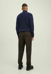 Uomo in piedi di spalle, con un maglione blu navy, pantaloni marrone scuro con tasche posteriori con bottoni e scarpe nere su uno sfondo neutro.