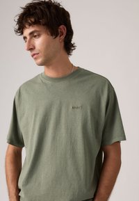 Νεαρός με σγουρά σκούρα μαλλιά που φοράει πράσινη μπλούζα Levi’s και ασημένιο αλυσίδα στον λαιμό, κοιτάζοντας προς τα κάτω και αριστερά του.