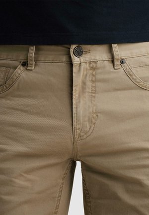 Záber zblízka na béžové nohavice s čiernym gombíkom a detailmi švov, nosené s čiernou košeľou zastrčenou dovnútra.