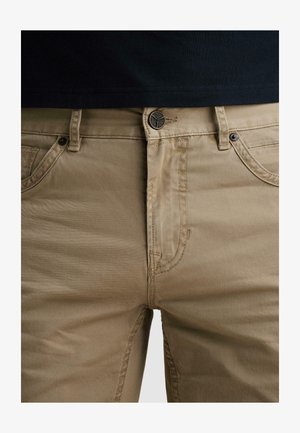 Nahaufnahme von beigen Hosen mit schwarzem Knopf und Steppnähten, getragen mit einem in die Hose gesteckten schwarzen Hemd.