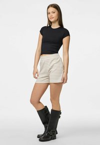 Crna prilagodljiva majica, bež šortsevi s bijelim prugama i crne kožne čizme do koljena s kopčama i teksturiranom završnom obradom.