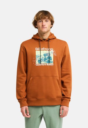 Giovane uomo che indossa una felpa con cappuccio marrone Billabong con tasca frontale e stampa grafica, abbinata a pantaloni verde chiaro, in piedi su sfondo bianco.