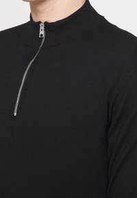 Pullover nero con colletto alto, caratterizzato da un dettaglio con zip. Realizzato in materiale testurizzato, presenta un design aderente.