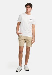 Hvit kortermet t-skjorte med en liten svart logo, kombinert med beige shorts og grå-og-hvite joggesko. Enkelt, casual antrekk.