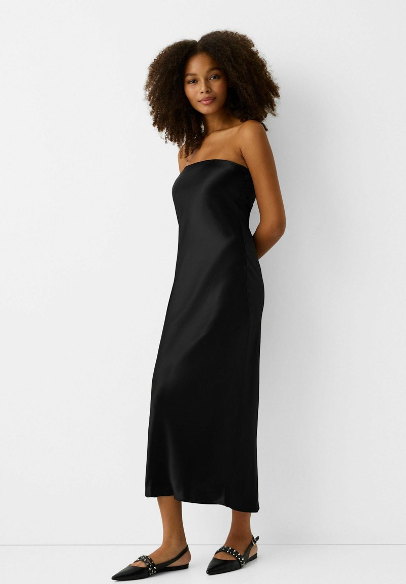 Zwarte satijnen strapless jurk met een gladde textuur, midi-lengte en een aansluitende vorm. Gecombineerd met zwarte puntige ballerina's met studaccenten.