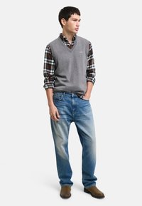 Grå ullblandad ärmlös tröja över en rutmönstrad skjorta med knappar, ihop med ljusblå denimjeans och bruna mockakängor.