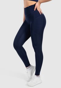 Leggings taille haute bleu marine avec une texture lisse, dotés de poches latérales et de coutures renforcées le long des coutures, associés à des chaussures de sport blanches.