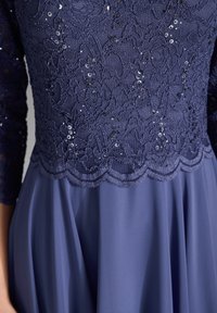 Navyblaues Kleid mit einem Spitzenoberteil, bestickt mit Pailletten, mit gewelltem Detail und einem fließenden Satinsaum. Elegante und strukturierte Stoffkombination.