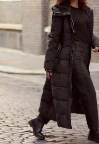 Langer schwarzer Steppmantel mit Kapuze, Rautenmuster und Reißverschlussdetails, kombiniert mit schwarzen weitem Hosenbeinen und schwarzen Stiefeletten.