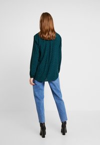 Blusa verde con estampado, de mangas largas, ligeramente holgada y con dobladillo redondeado, combinada con unos vaqueros de color azul claro y botas negras de tobillo.