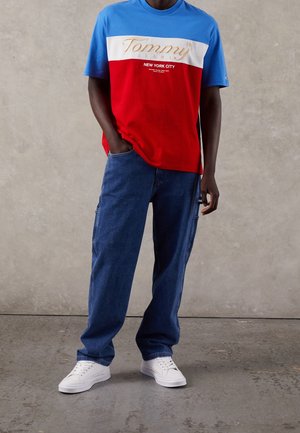 Camiseta color bloque en rojo y azul que presenta el logo "Tommy Jeans" en dorado, combinada con unos jeans azules de corte relajado y zapatillas blancas.