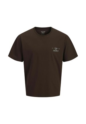 Garçon debout et souriant, portant un t-shirt marron foncé, un jean bleu clair et des chaussures beiges à lacets sur un fond clair uni.