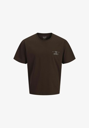 Garçon debout et souriant, portant un t-shirt marron foncé, un jean bleu clair et des chaussures beiges à lacets sur un fond clair uni.