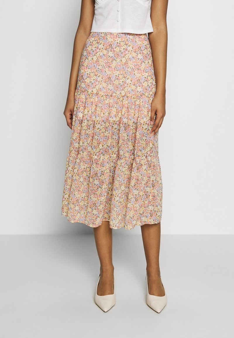 Jupe midi florale dans des couleurs pastel, présentant un design à volants, un tissu doux et une silhouette évasée. Associée à des escarpins beiges à bout pointu.