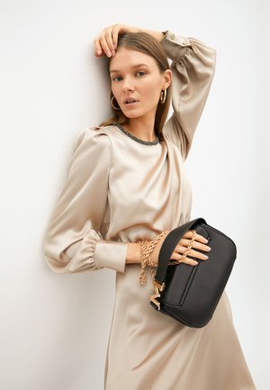 Borsa a tracolla nera con finitura in pelle liscia, accentuata da una tracolla in catena dorata e hardware geometrico, abbinata a un abito beige in satin.