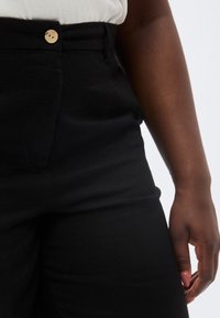 Κοντινό πλάνο ατόμου που φοράει μαύρο παντελόνι με ξύλινο κουμπί και λευκό πουκάμισο μαζεμένο μέσα, δείχνοντας μέρος από το δεξί του χέρι και χέρι.